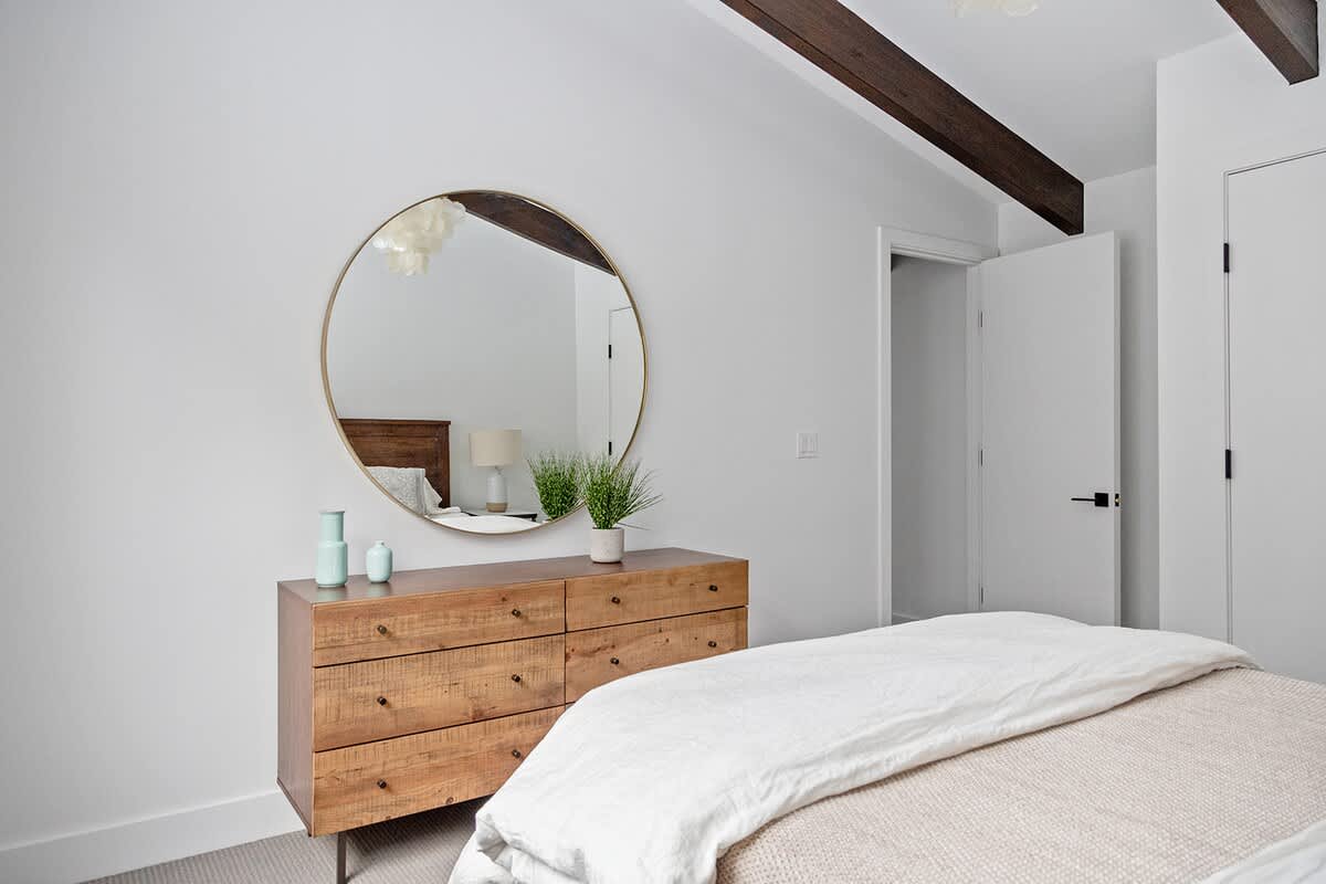 Bedroom view highlighting a rustic wooden dresser with a large round mirror, reflecting the room's light. Exposed dark wooden beams on the ceiling add architectural interest.