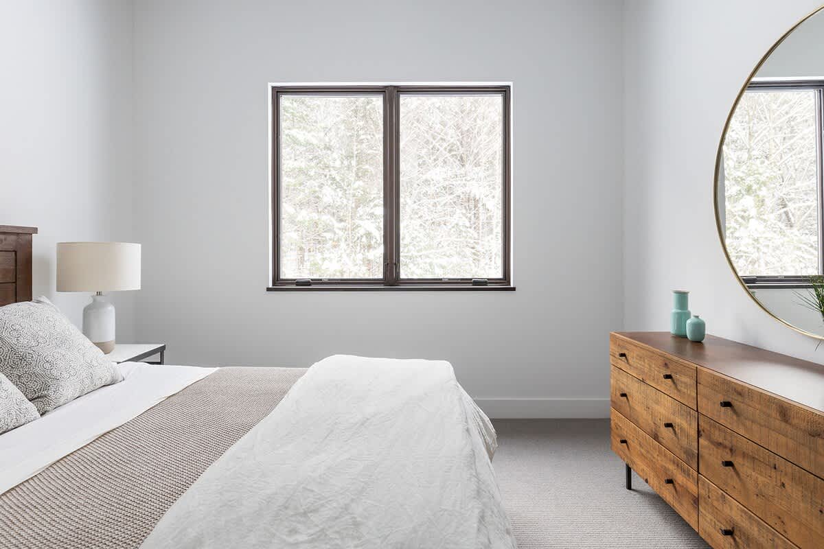 View of a bedroom featuring a large window with a snowy outdoor scene, a wooden bed, and a rustic wooden dresser topped with a round mirror. White walls and light gray carpet complete the space.