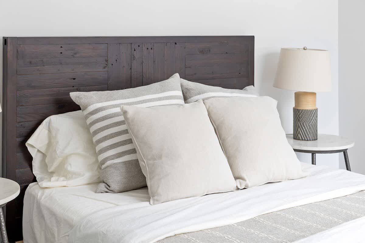 Close-up of a bed featuring a dark wooden headboard, white sheets, and a mix of striped gray and solid beige throw pillows. A bedside lamp is partially visible on the right.