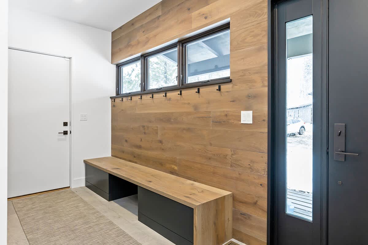 A modern entryway features a long wooden bench with dark drawers, a wood-paneled accent wall with coat hooks, and large windows, providing a functional and stylish welcome.