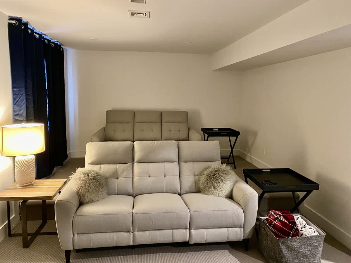 A cozy media room with two rows of light-colored reclining sofas, each with three seats, and two black tray tables, set on a light carpet with white walls.