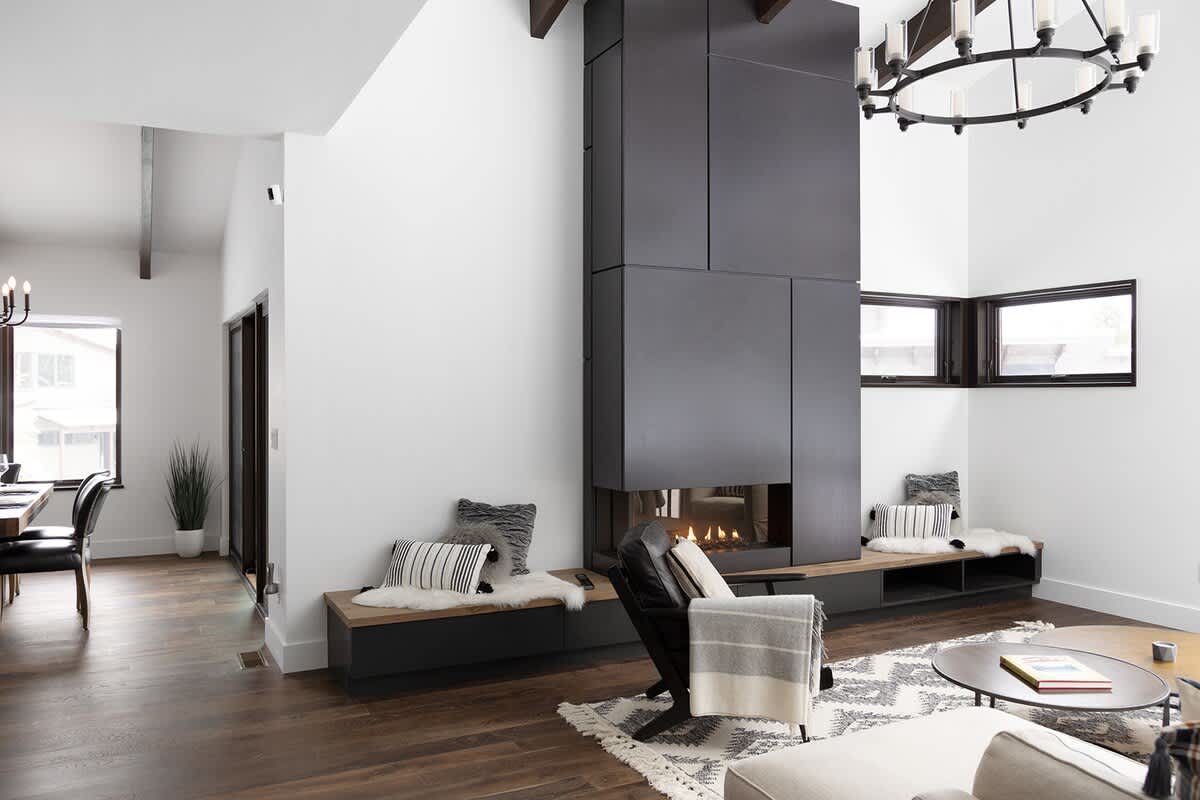 A modern open-concept living space features a striking black fireplace column, a comfortable seating area with a patterned rug, and a glimpse of a dining area in the background, all with hardwood floors.