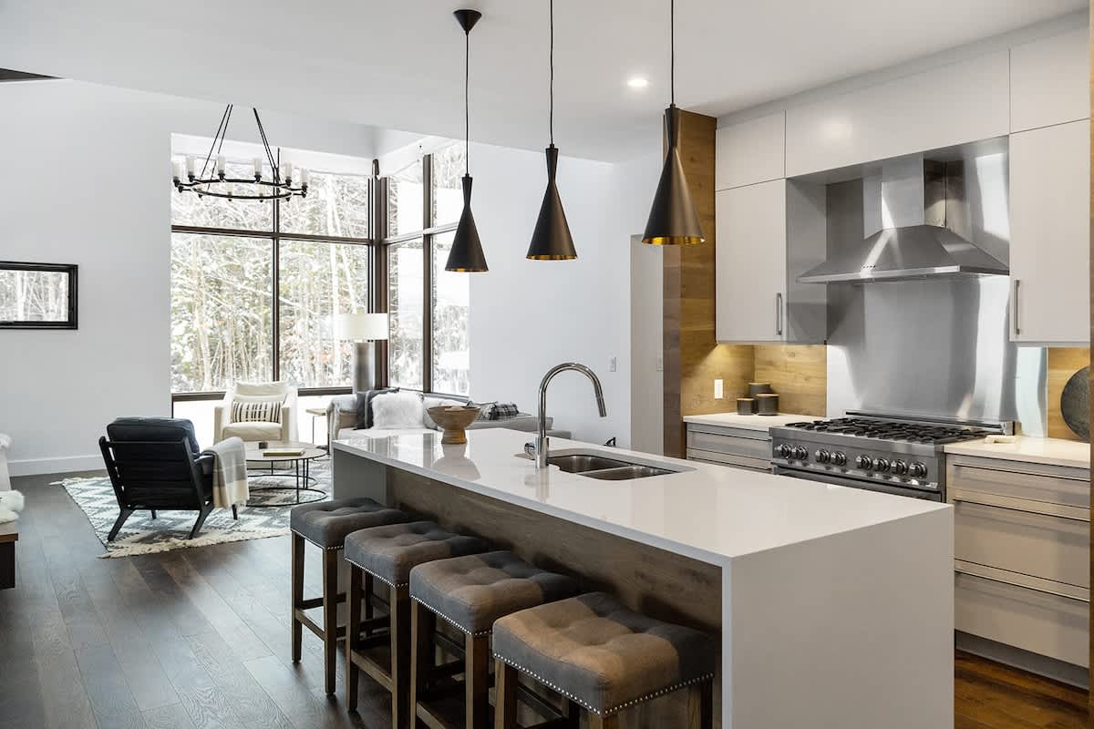 Modern open-concept kitchen with a large white island, dark gray bar stools, stainless steel appliances, and dark pendant lights, overlooking a living area with large windows and snowy trees outside.