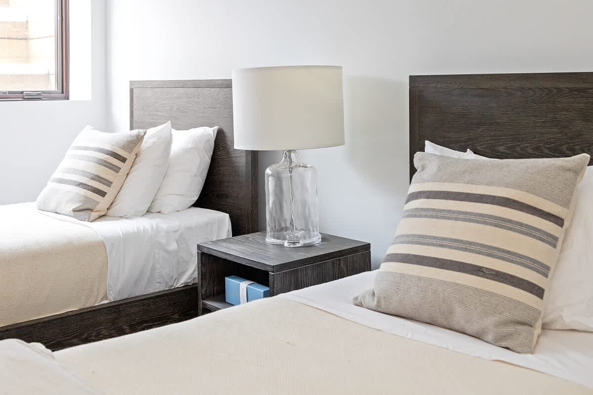 A bright bedroom featuring two twin beds with dark wood frames, white bedding, and striped throw pillows, separated by a dark wood nightstand with a clear glass lamp.