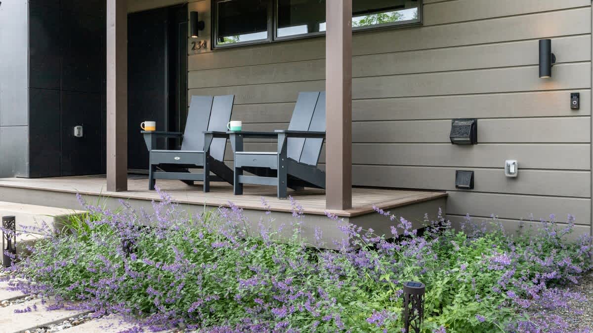 The front porch of a modern house with gray siding, featuring two dark Adirondack chairs with mugs on a small table, surrounded by lush purple flowering plants.