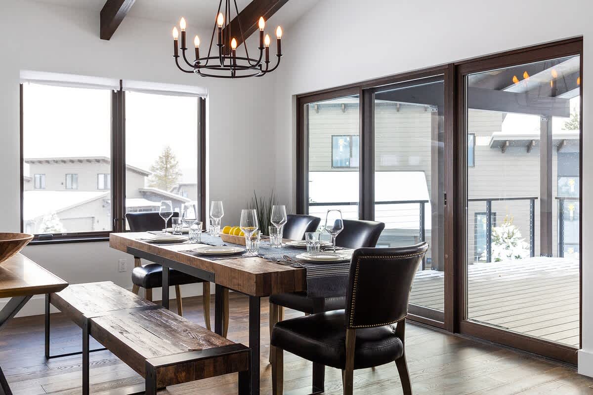 Spacious dining room with a long wooden table, black leather chairs, a matching wooden bench, and a black chandelier, surrounded by large windows and sliding glass doors offering views of a snowy landscape.