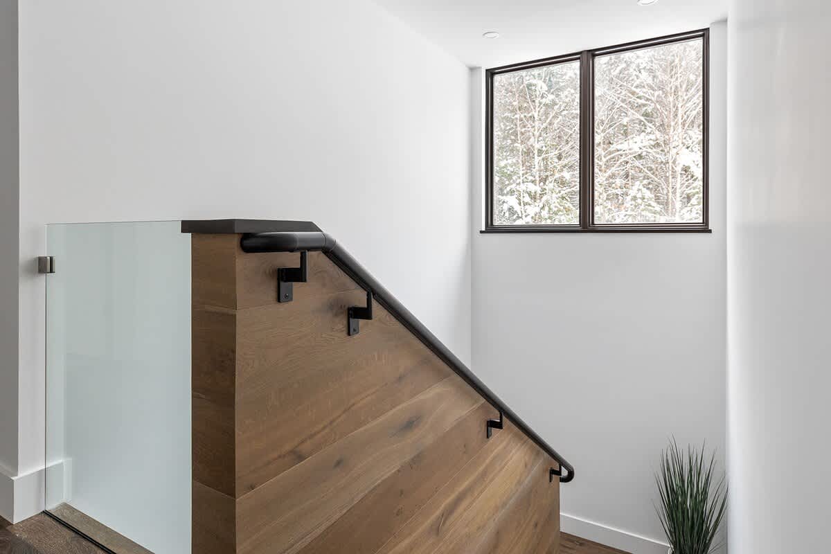Modern staircase with a wooden base, black handrail, and glass panel railing, leading up to a landing with a large window offering a view of snowy trees.
