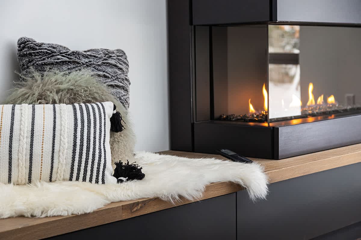 Close-up of a modern gas fireplace with a glass enclosure, set into a black structure with a wooden hearth, adorned with decorative pillows and a white faux fur throw.