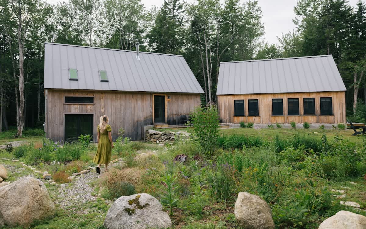Two modern rustic cabins with dark wood siding and gray metal roofs are set in a natural landscape with large rocks, a gravel path, and wild grasses. A woman in a green dress walks away from the camera.