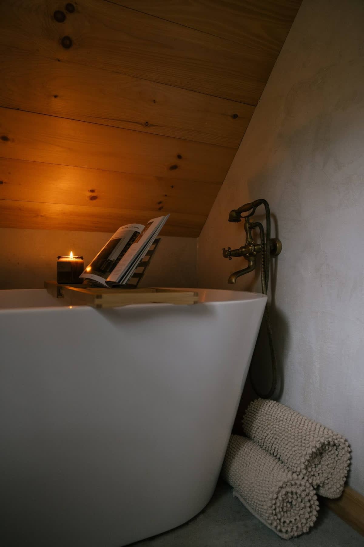 A freestanding white bathtub is nestled under a sloped wooden ceiling, illuminated by a warm candle on a bath caddy with an open book. Vintage brass fixtures are mounted on the wall, and rolled towels are on the floor.