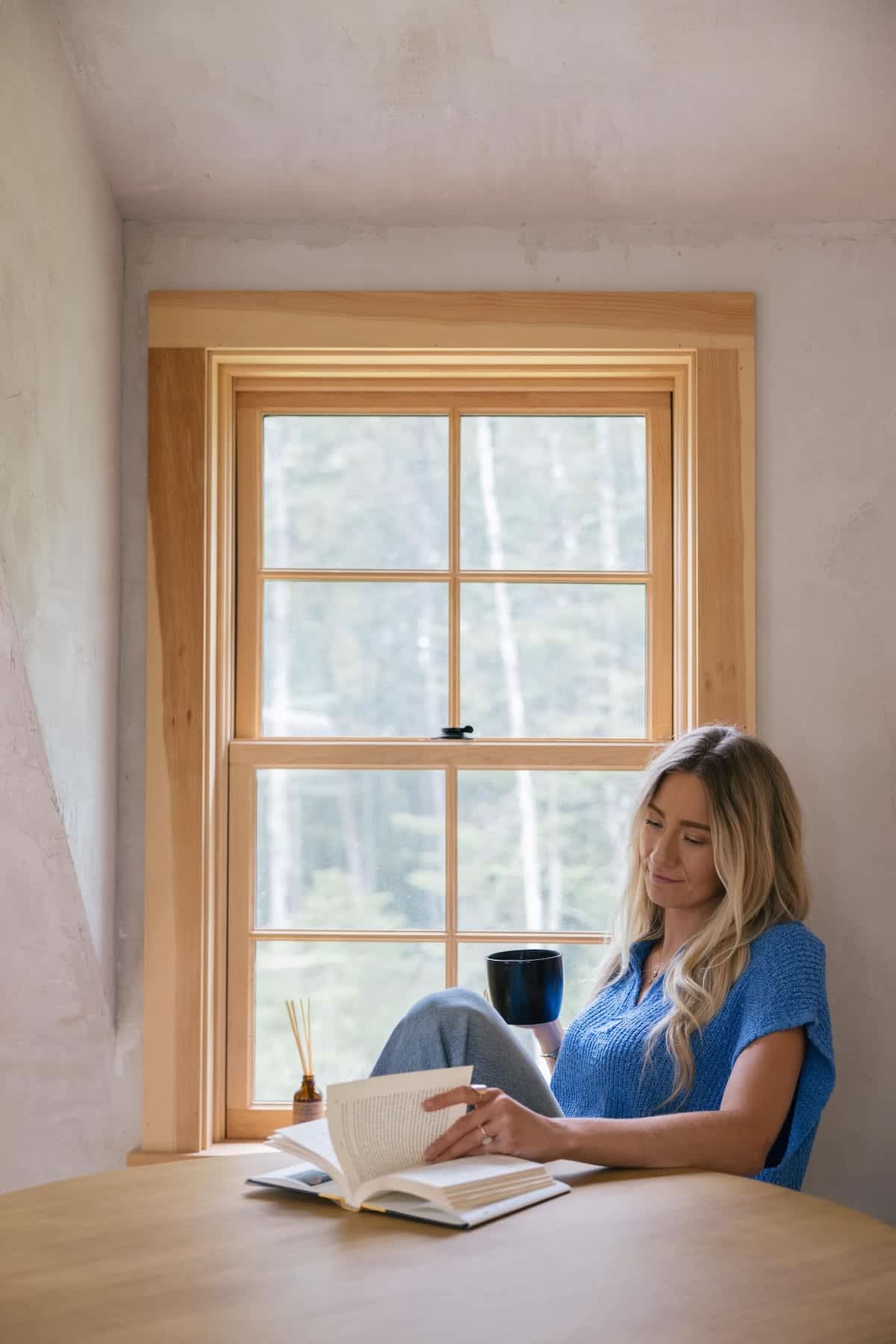 A woman with blonde hair reads a book while sitting comfortably by a large window with wooden trim, holding a dark mug. A diffuser and another book are on a light wooden table in front of her.
