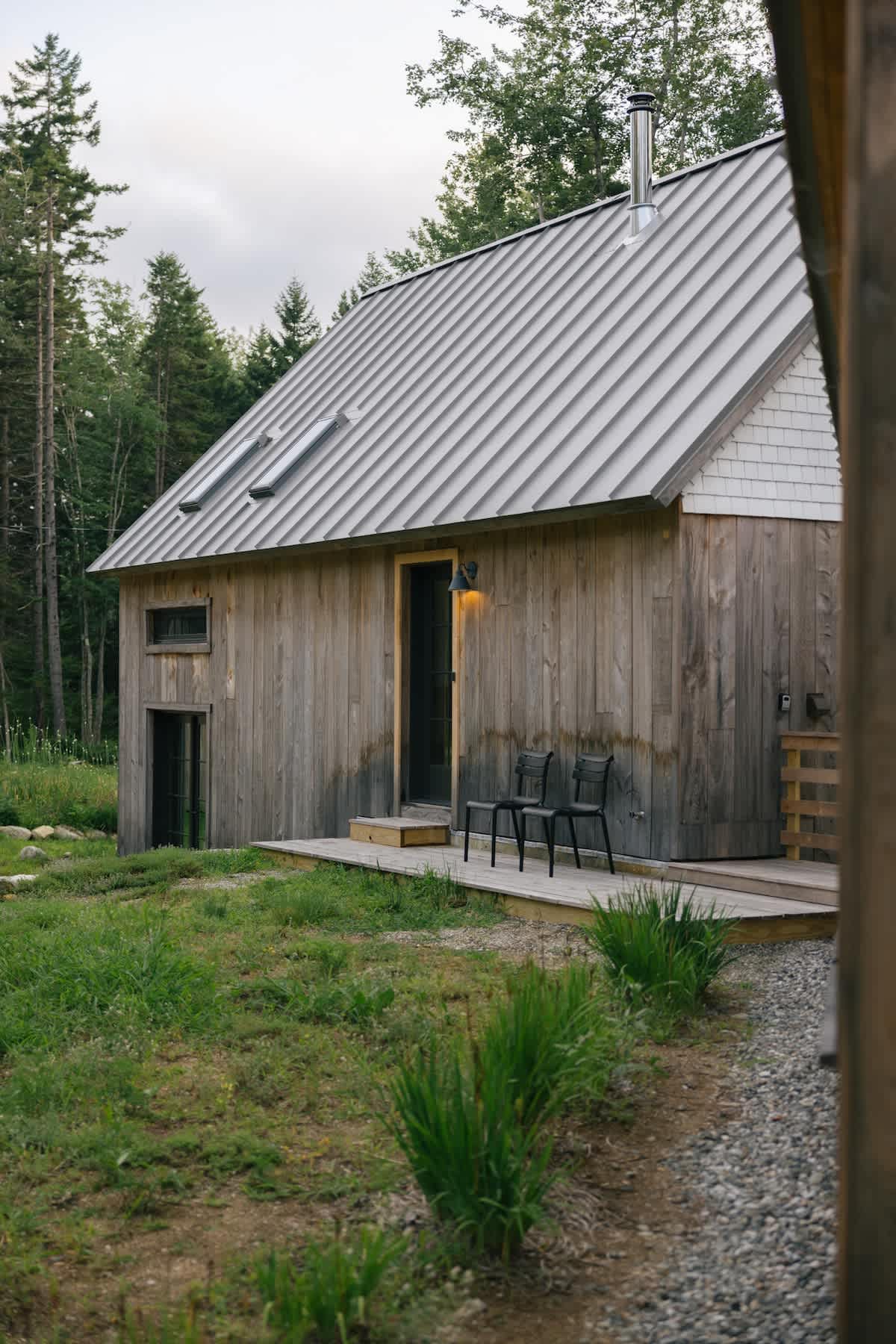 A close-up view of a modern cabin with natural wood siding and a light metal roof, featuring a small wooden deck with two black chairs. Lush green grass and trees surround the building.