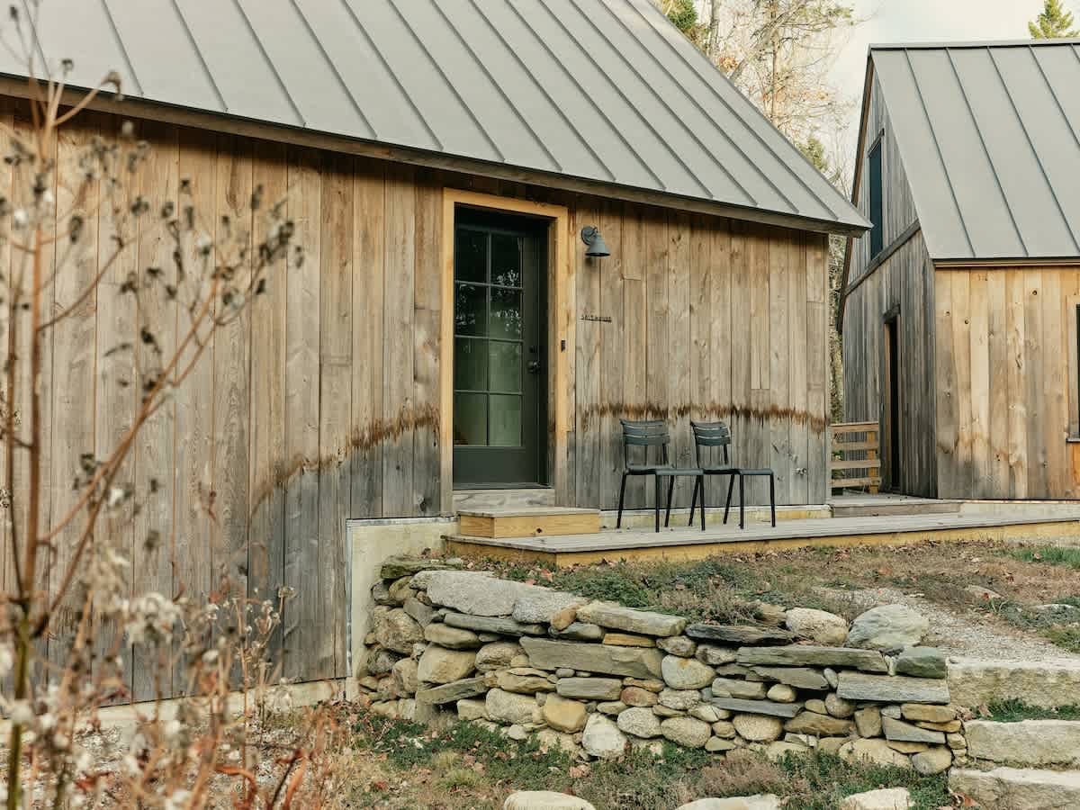 A close-up of a rustic wooden cabin's entrance, featuring a dark-framed glass door and a small wooden deck with two chairs. A low dry-stacked stone wall borders the foreground, with wild vegetation.