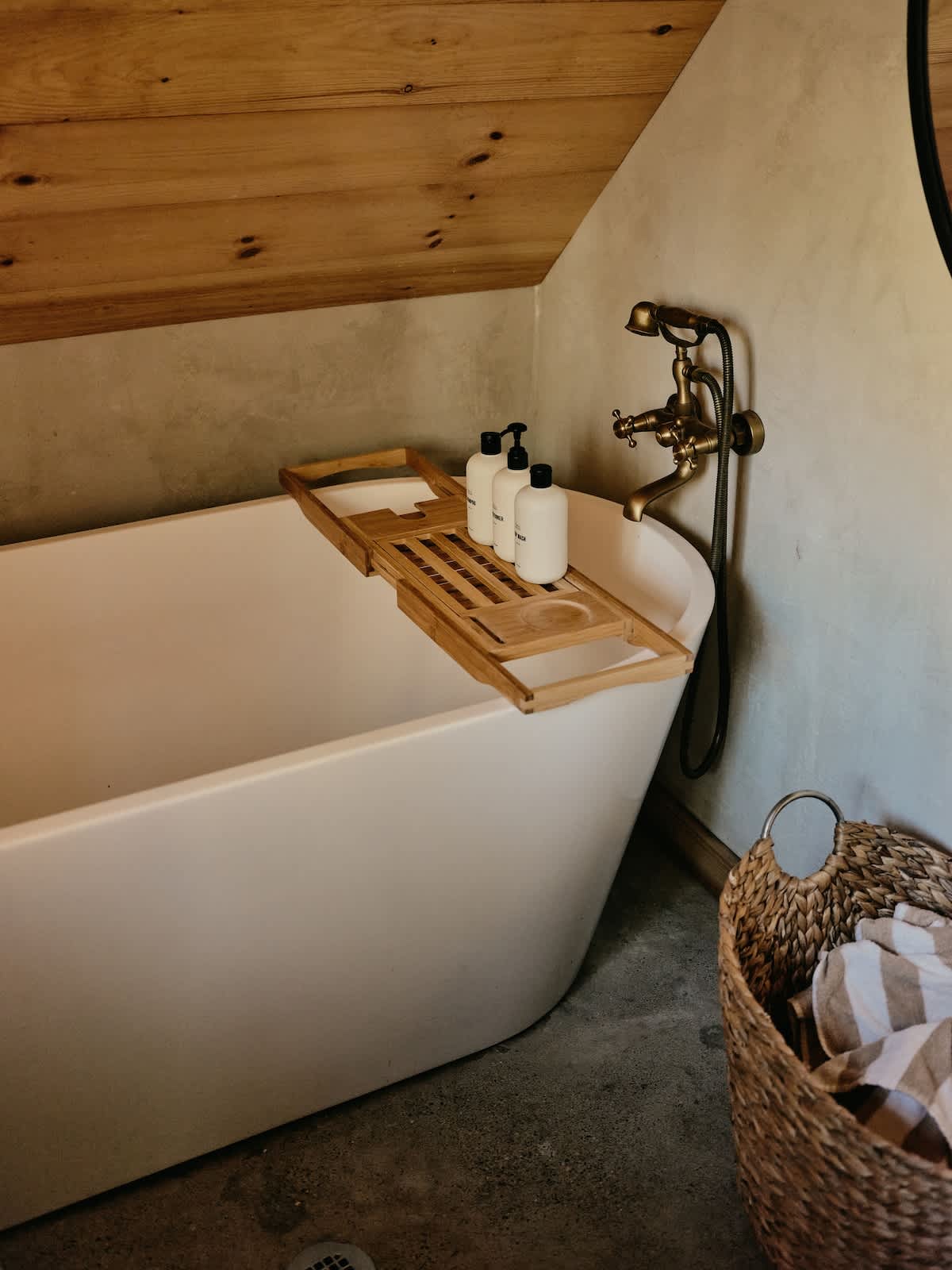 A white freestanding bathtub features a wooden bath caddy holding three white bottles of bath products. Vintage brass fixtures are mounted on the wall, and a woven basket with towels is next to the tub.