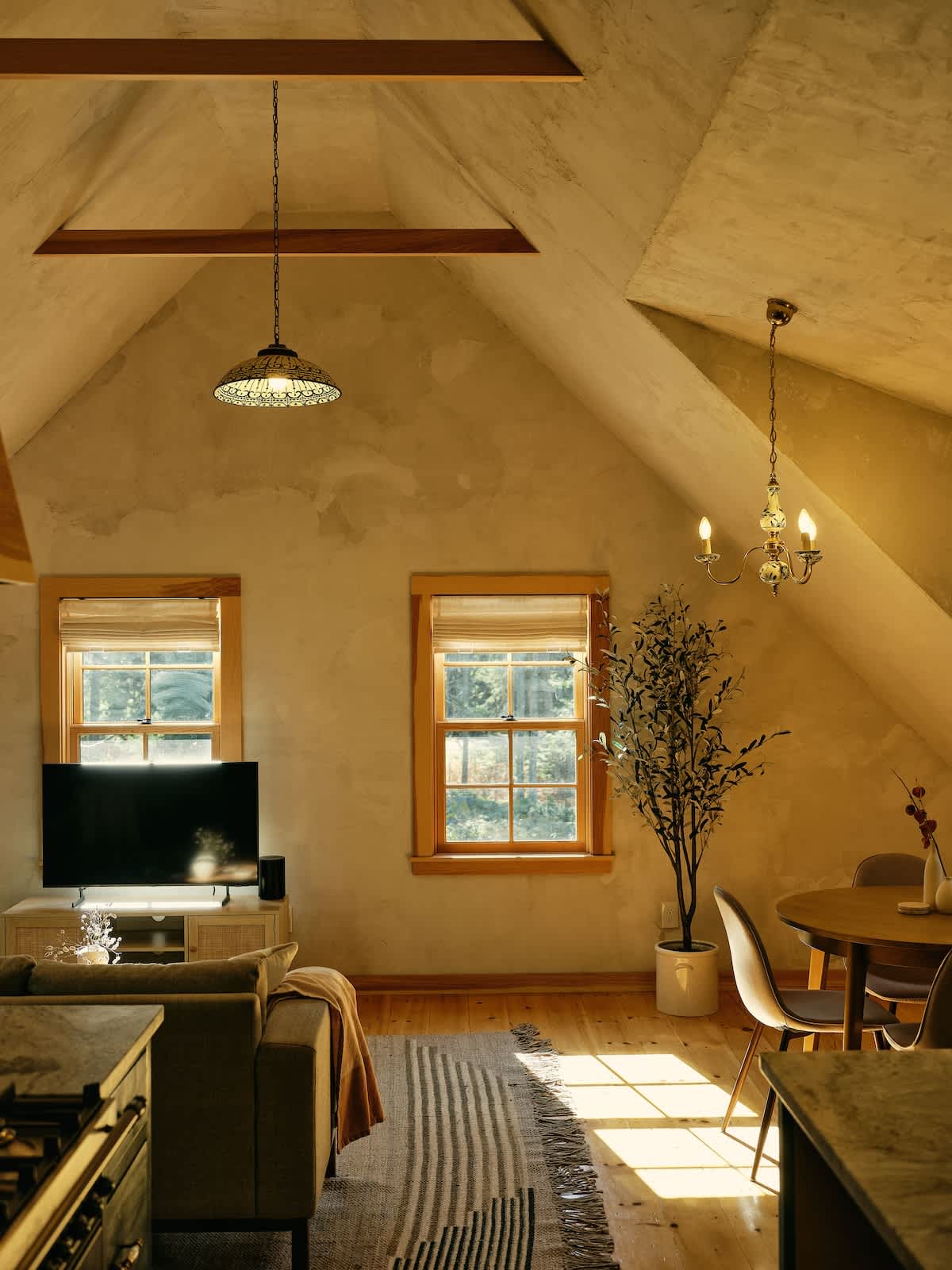 A spacious open-concept living area features a sofa, a TV on a console, and a dining table with chairs, all illuminated by natural light from multiple windows and overhead pendant lights, with a vaulted ceiling.