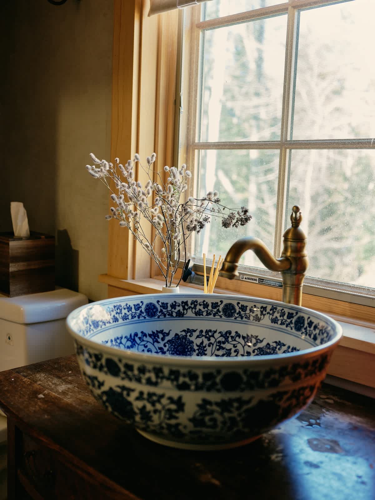 A decorative blue and white ceramic bowl sink sits on a rustic wooden vanity next to a window, with a vintage brass faucet. Dried flowers in a small vase and a tissue box are also visible.
