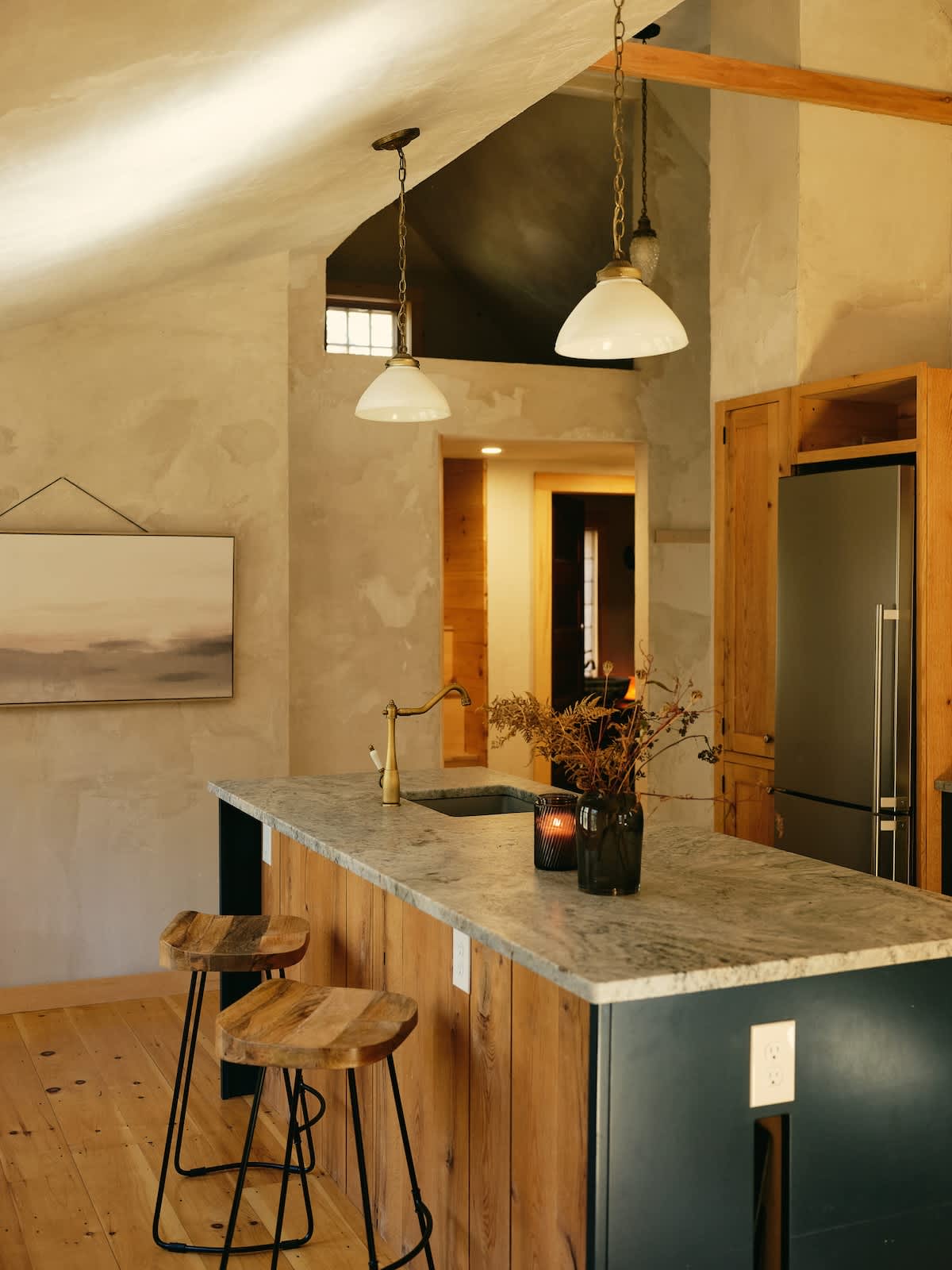 The kitchen island, clad in vertical wood planks and topped with light gray stone, features a brass faucet and sink, with two wooden bar stools and two white pendant lights hanging from the vaulted ceiling.