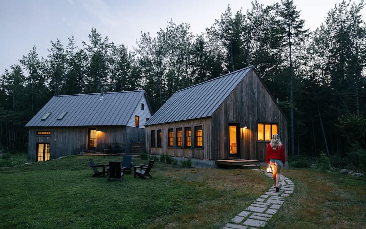 Two modern rustic cabins with dark wood siding and gray metal roofs stand amidst a grassy yard and tall evergreen trees at dusk. A person walks on a stone path, carrying a lantern, towards the illuminated windows.
