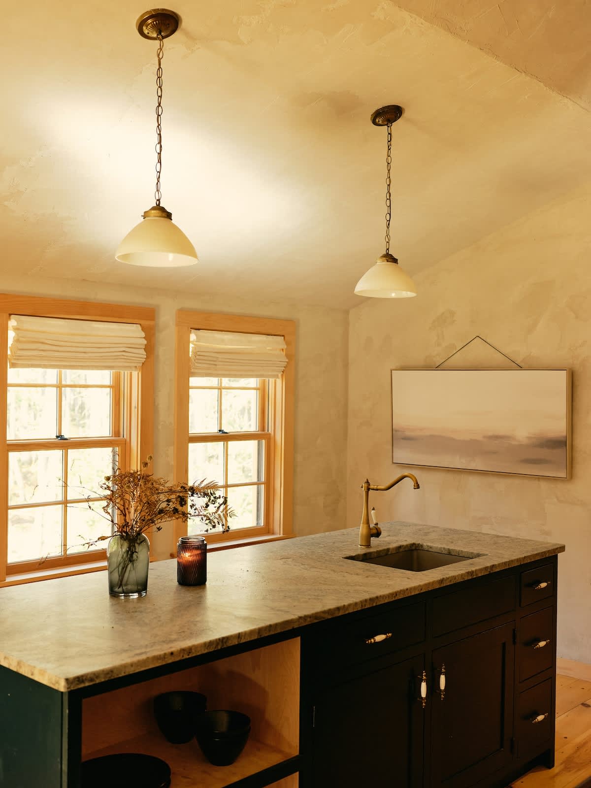 A warm-toned kitchen features a dark island with a sink and brass faucet, illuminated by two pendant lights. Two windows with light-colored Roman shades flank the textured, plaster-like walls.