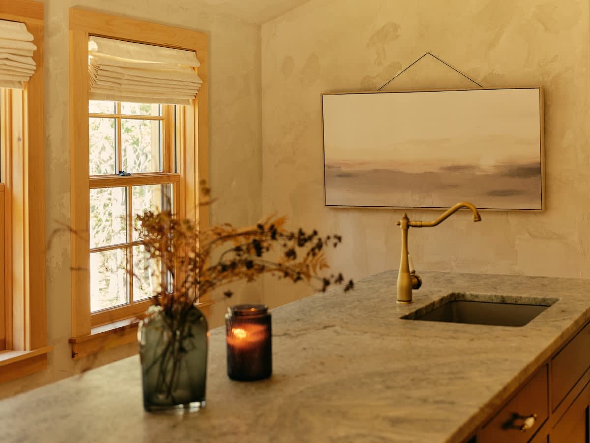 A close-up of the kitchen island showcases a light-colored, marble-patterned countertop with a brass faucet and sink. A vase of dried plants and a lit candle add a cozy touch, with windows in the background.