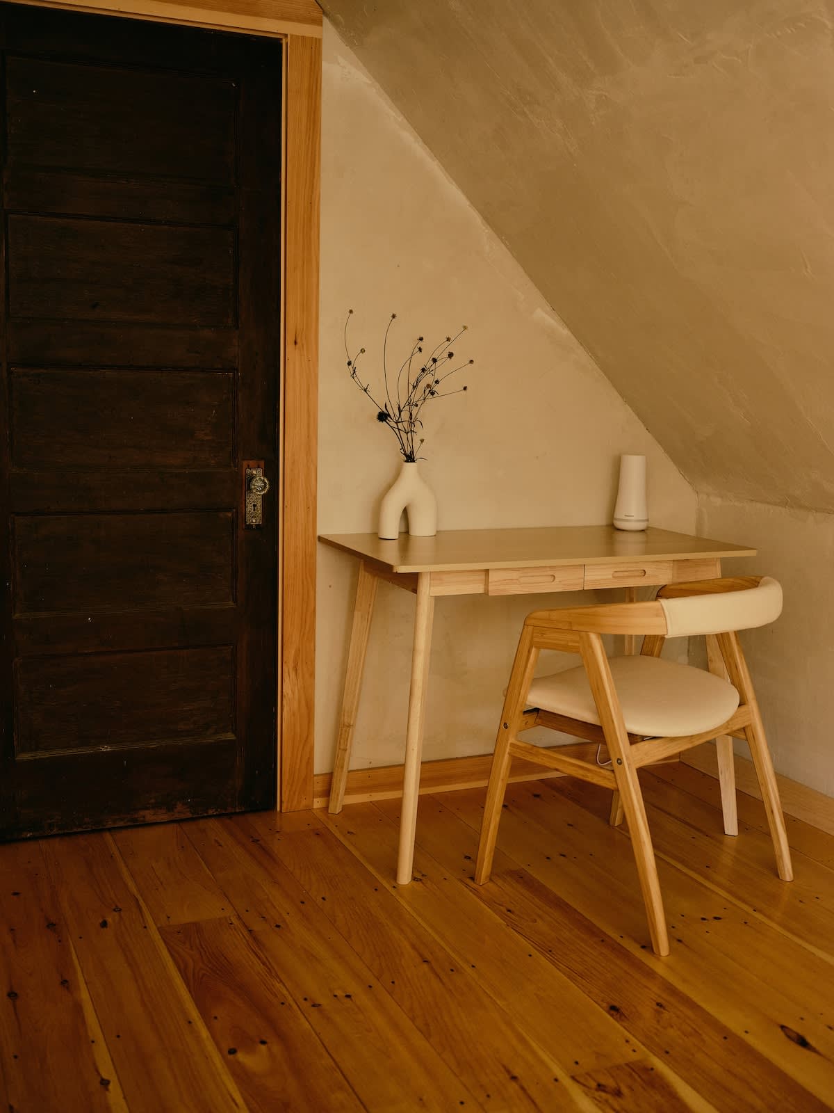 A light-toned wooden desk with a modern white vase and dried flowers sits against a textured light wall, next to a dark wooden door. A matching wooden chair with a light cushion is positioned in front of the desk, all on warm hardwood floors.
