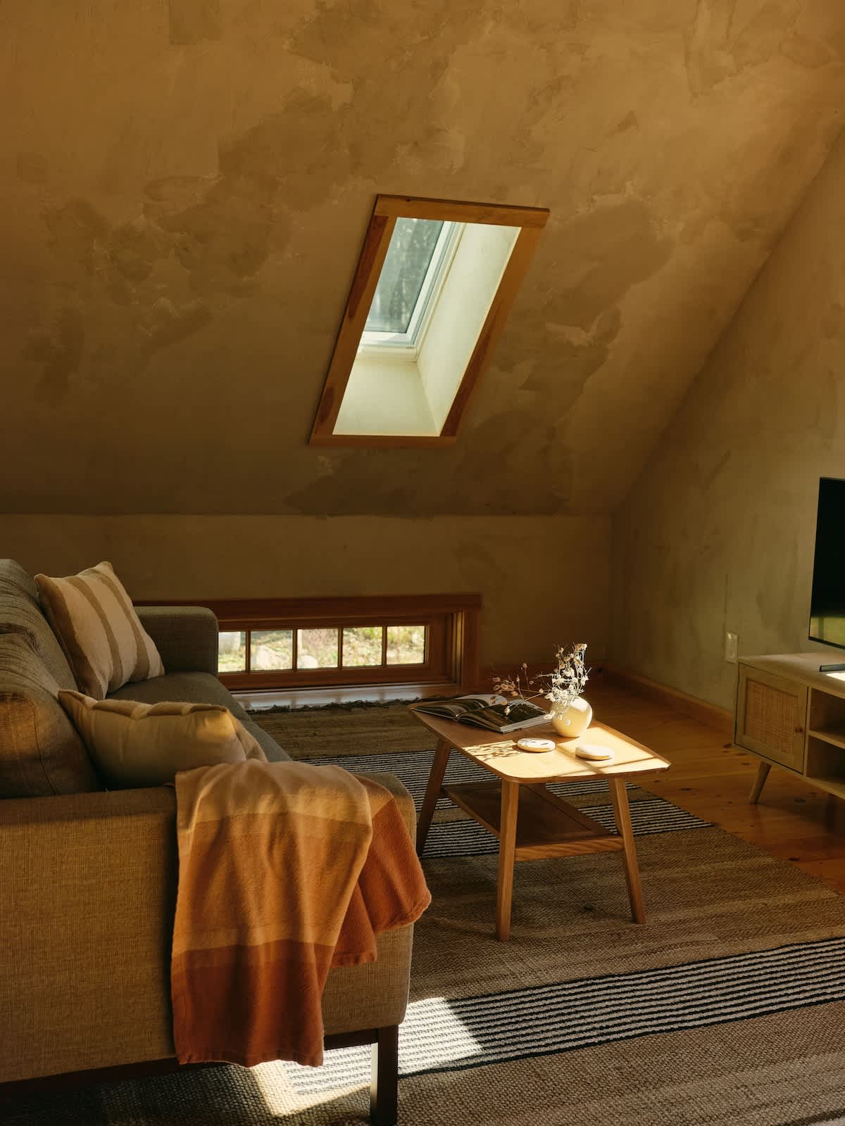 A cozy living room features a light gray sofa, a wooden coffee table, and a striped rug on hardwood floors. The sloped ceiling includes a rectangular skylight, bathing the room in warm sunlight.