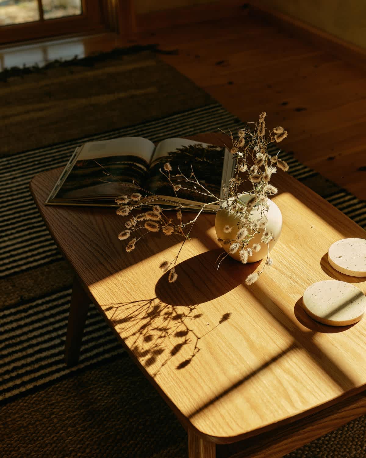 A close-up of a wooden coffee table displays an open book, a white vase with dried flowers, and two round coasters. Sunlight streams in, casting intricate shadows across the table and the striped rug.