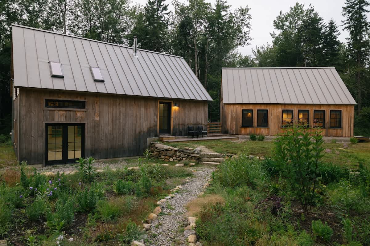 Two modern cabins with natural wood siding and light metal roofs are set amidst a lush green landscape with gravel paths and wild vegetation. The larger cabin has a small deck with outdoor chairs.