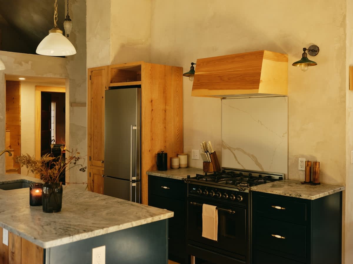 A rustic-modern kitchen features dark lower cabinets with a gas range, a stainless steel refrigerator integrated into light wood cabinetry, and a light-colored stone countertop with a matching backsplash behind the stove.