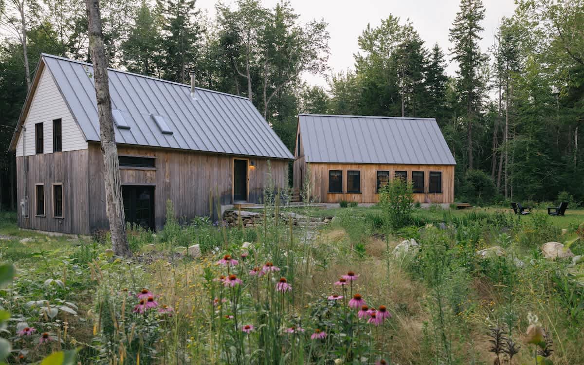 Two modern rustic cabins with dark wood siding and gray metal roofs are nestled in a naturalized garden with pink coneflowers and tall trees. The larger cabin has skylights and a small deck.