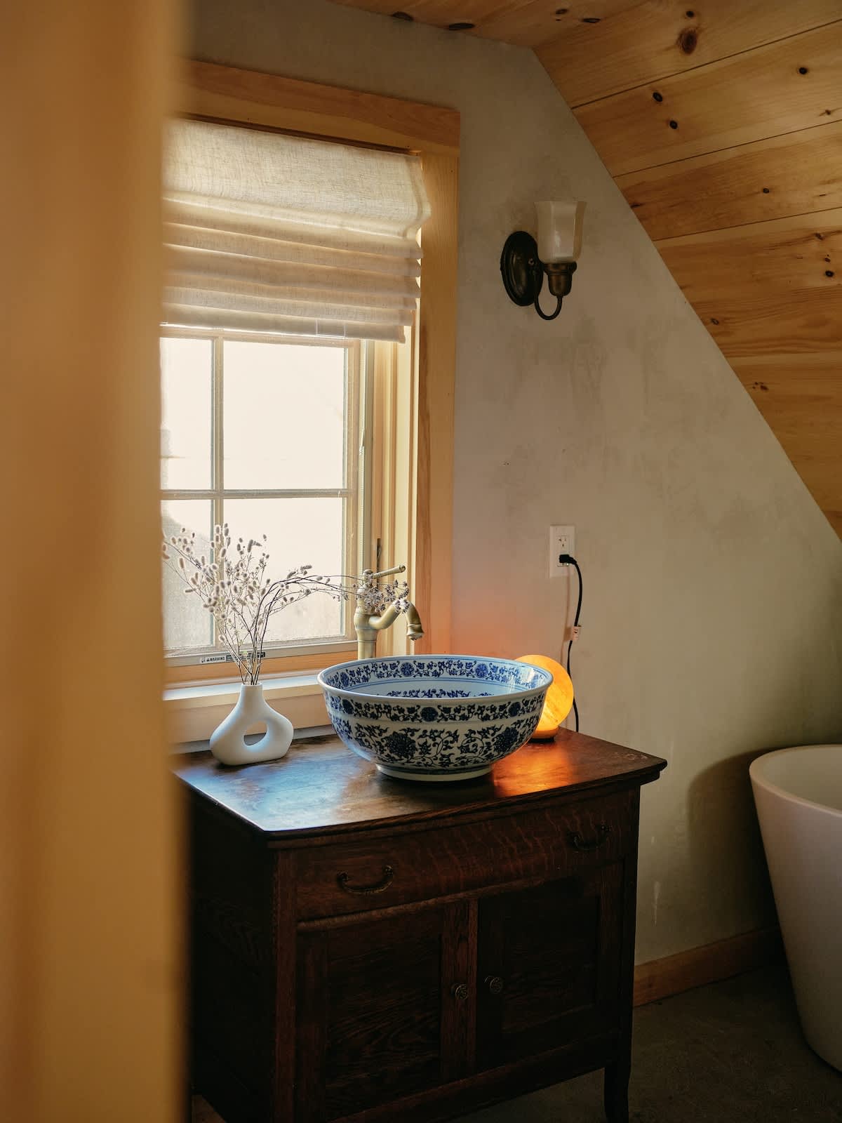 A rustic wooden vanity with a blue and white ceramic bowl sink and vintage brass faucet is positioned under a window with a roman shade. Dried flowers in a vase and a wall sconce are visible, with a white bathtub partially seen on the right.