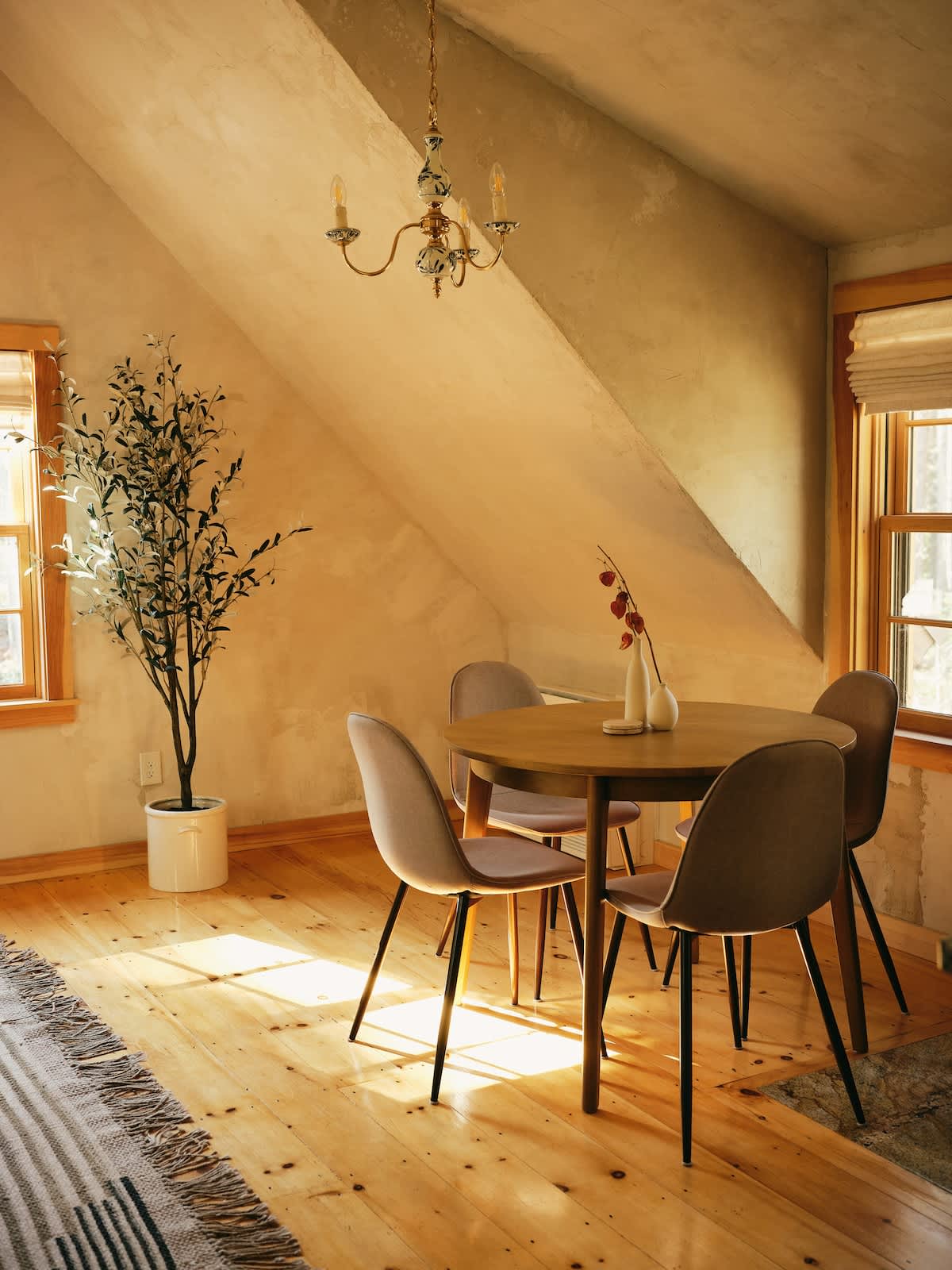 A bright dining area features a round wooden table surrounded by four modern chairs with light-colored upholstery. A tall faux olive tree stands in a white pot, and a vintage-style chandelier hangs from the sloped ceiling.