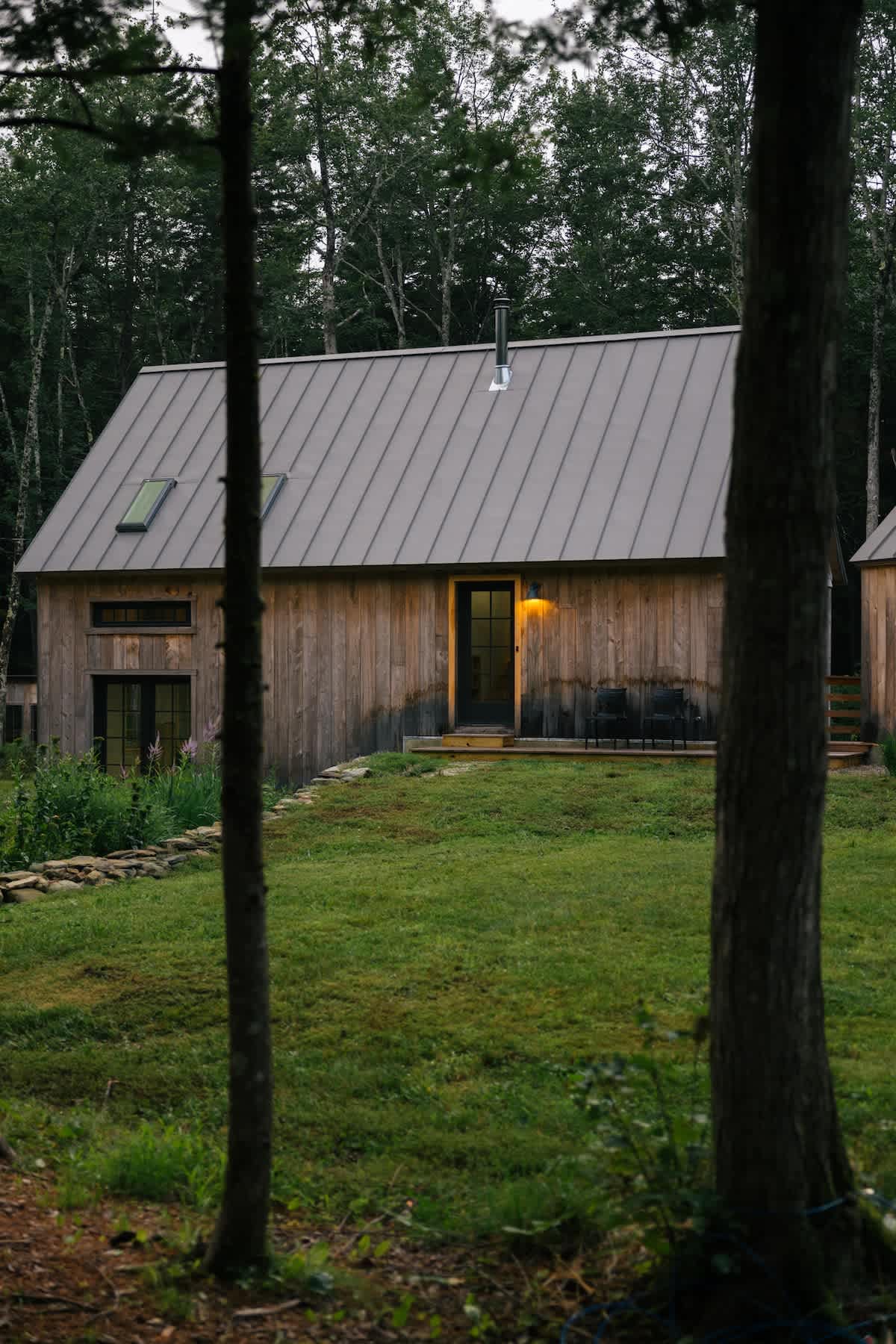 A rustic wooden cabin with a gray metal roof and skylights features an illuminated black front door with a small porch and two black outdoor chairs. Tall trees partially obscure the view.