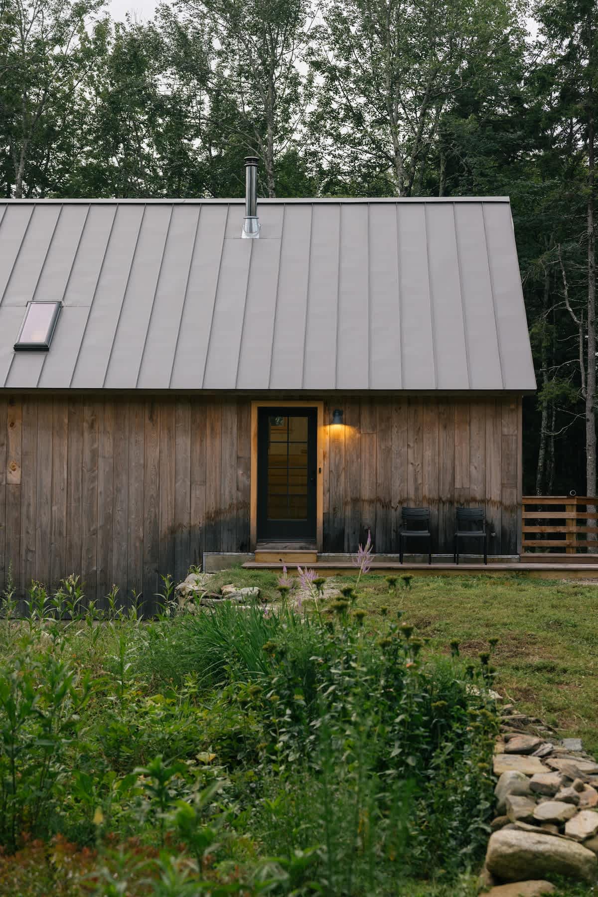 The front exterior of a rustic barn-style house with vertical wooden siding and a gray metal roof, featuring a dark door, an outdoor light, and a chimney, surrounded by green grass and tall trees.