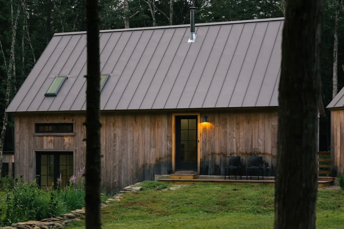 A rustic wooden cabin with a gray metal roof and skylights features an illuminated black front door with a small porch and two black outdoor chairs. Tall trees partially obscure the view.