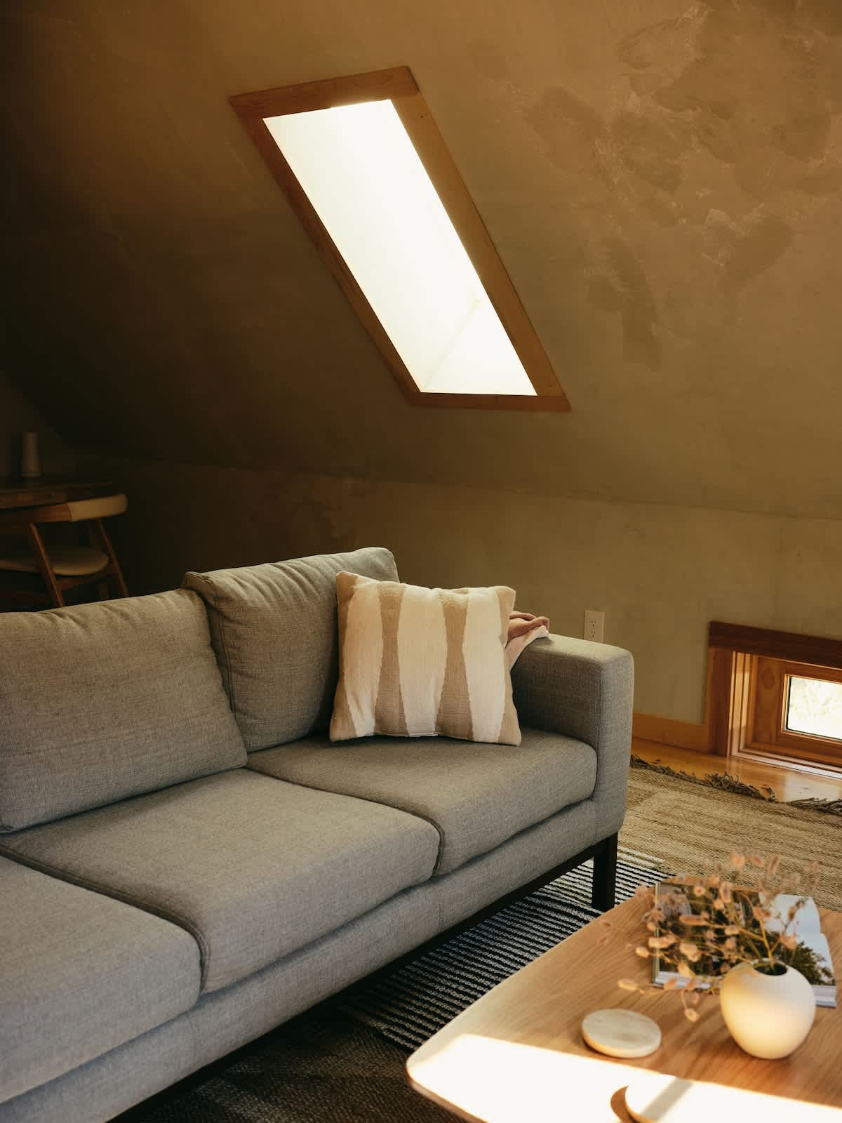 A comfortable light gray sofa with a striped throw pillow and a blanket is positioned in a living room with a sloped ceiling. A rectangular skylight and a small window provide natural illumination.