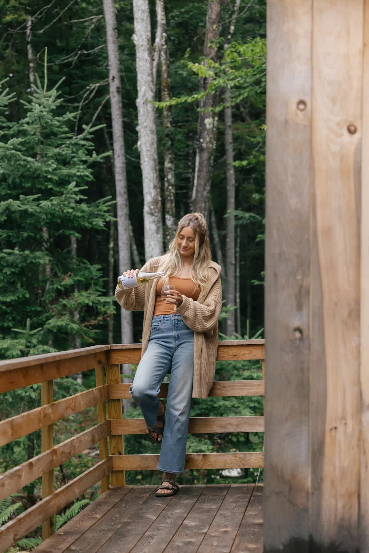 A woman with blonde hair pours wine into a glass on a rustic wooden deck, surrounded by tall green trees. She wears a tan cardigan, orange top, and jeans.