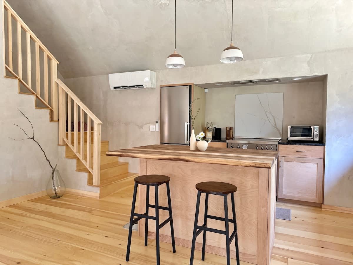 A modern kitchen with light wooden cabinetry and a large wooden island featuring a live-edge countertop and two dark bar stools. Stainless steel appliances, including a refrigerator and oven, are visible, along with two white pendant lights hanging above the island.
