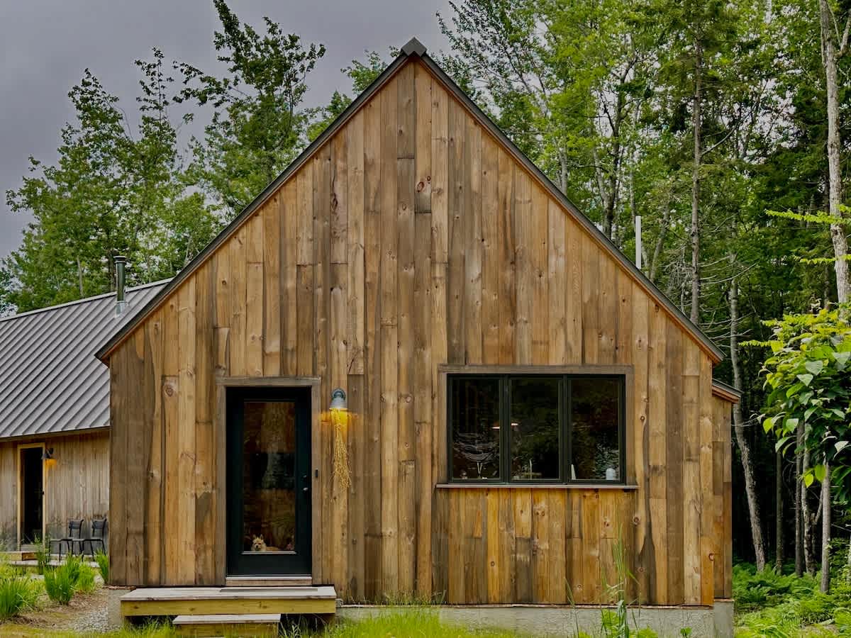 A rustic, gabled wooden cabin features vertical plank siding, a dark front door with a yellow wall sconce, and a large horizontal window, set against a backdrop of lush green trees.