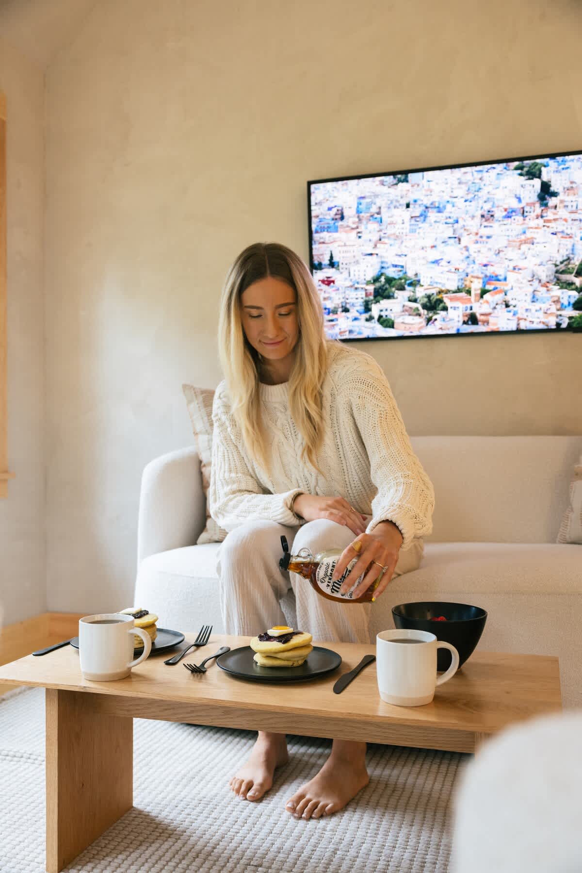 A cozy living room features a white sofa, a light wood coffee table, and a large wall-mounted TV displaying a colorful cityscape. A woman in a cream sweater enjoys pancakes and coffee, highlighting a relaxed atmosphere.