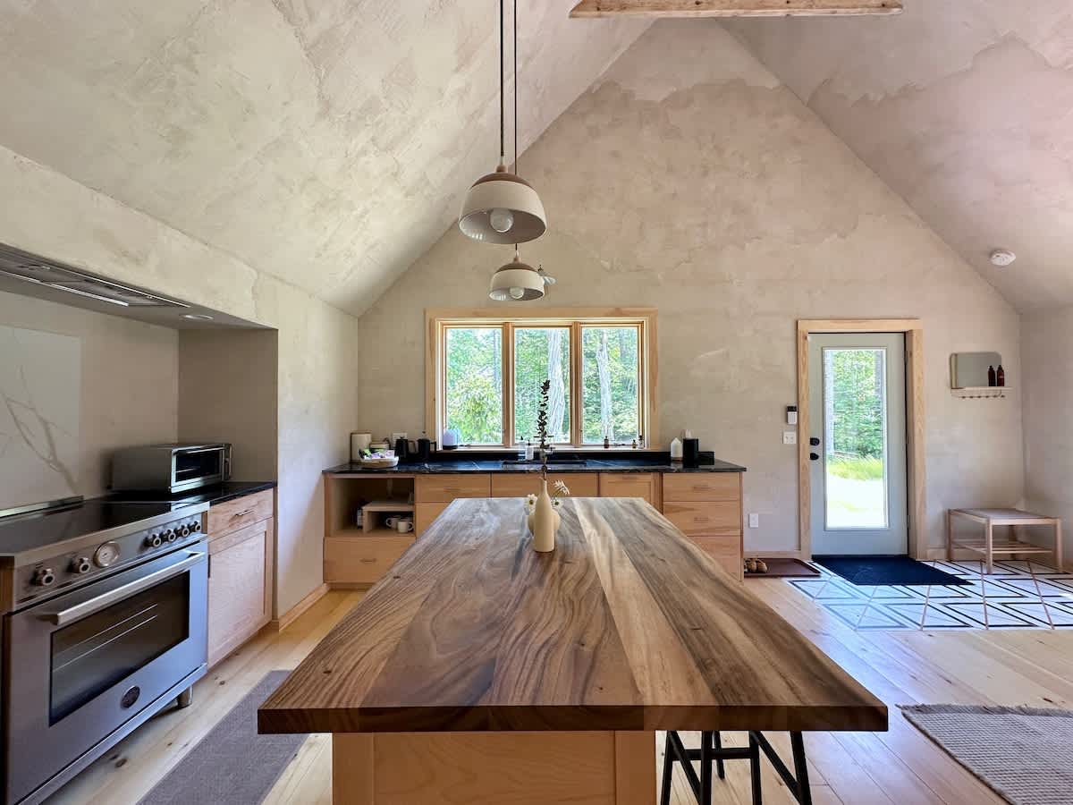Spacious kitchen with vaulted ceilings and light hardwood floors, featuring a large wooden island, light wood cabinetry, dark countertops, and a stainless steel range, with a window overlooking the forest.