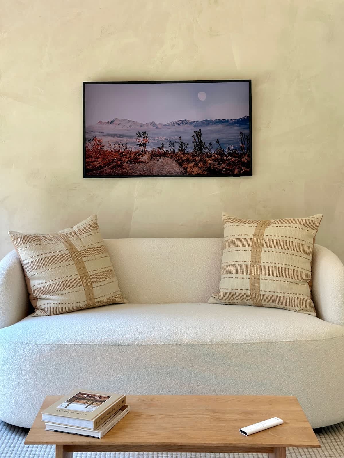 A minimalist living room features a curved white sofa with patterned throw pillows, a light wooden coffee table with books, and a large framed landscape artwork on a textured light-colored wall.