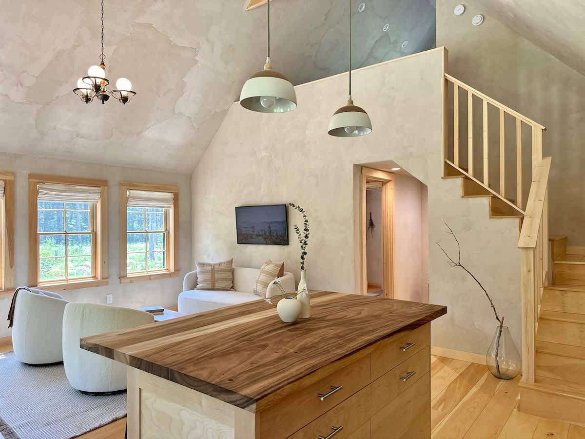 Open-concept living space with vaulted ceilings and light hardwood floors, featuring a comfortable seating area with a sofa and armchairs, and a large wooden kitchen island with pendant lights.