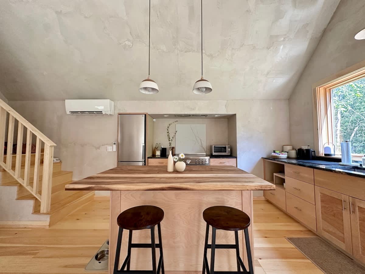 A spacious kitchen with light wooden floors and textured walls, centered around a large wooden island with a live-edge top and two bar stools. The kitchen features light wood cabinetry, stainless steel appliances, and a large window overlooking greenery.