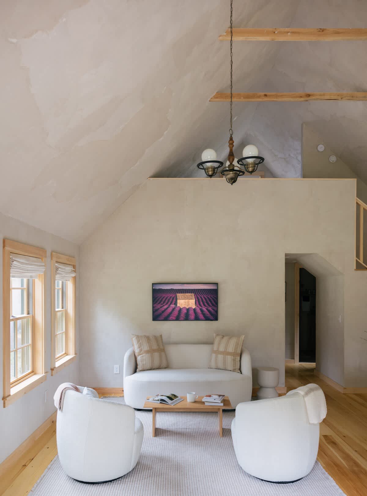 A spacious living room with a high vaulted ceiling and light textured walls, furnished with a cream sofa, two round white armchairs, and a wooden coffee table on an area rug. A wall-mounted TV displays a landscape image, and a wooden staircase is visible in the background.