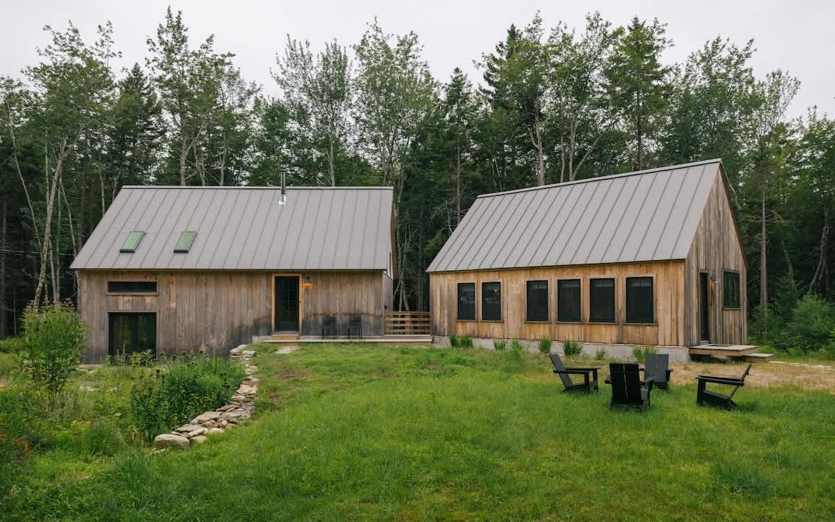 An exterior view of two modern wooden cabins with gray metal roofs, set in a grassy yard surrounded by tall trees. The cabins feature multiple windows and doors, with outdoor seating areas visible in the foreground.