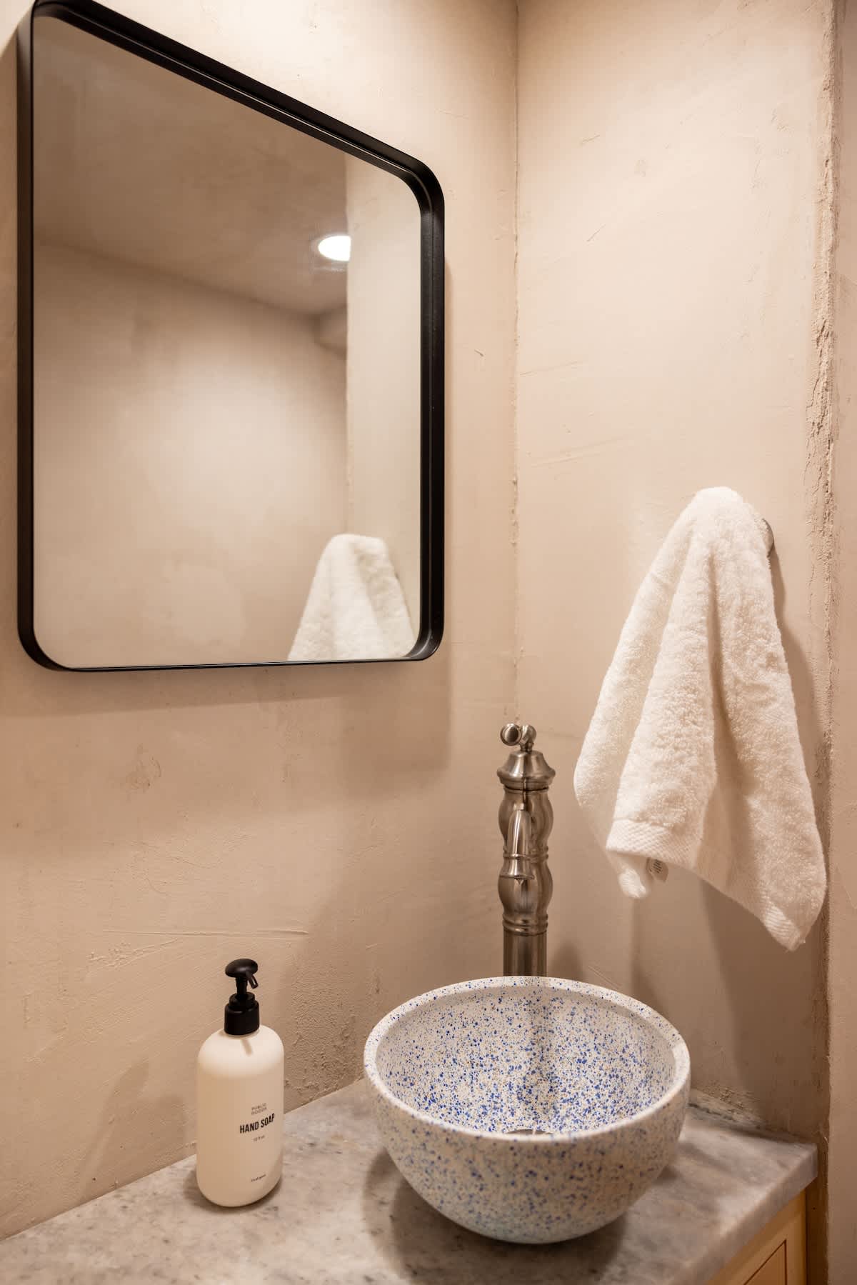 A modern bathroom with a black-framed rectangular mirror above a unique speckled blue and white bowl sink, a vintage-style silver faucet, and a white hand towel hanging on the wall. The walls are a light, textured plaster.