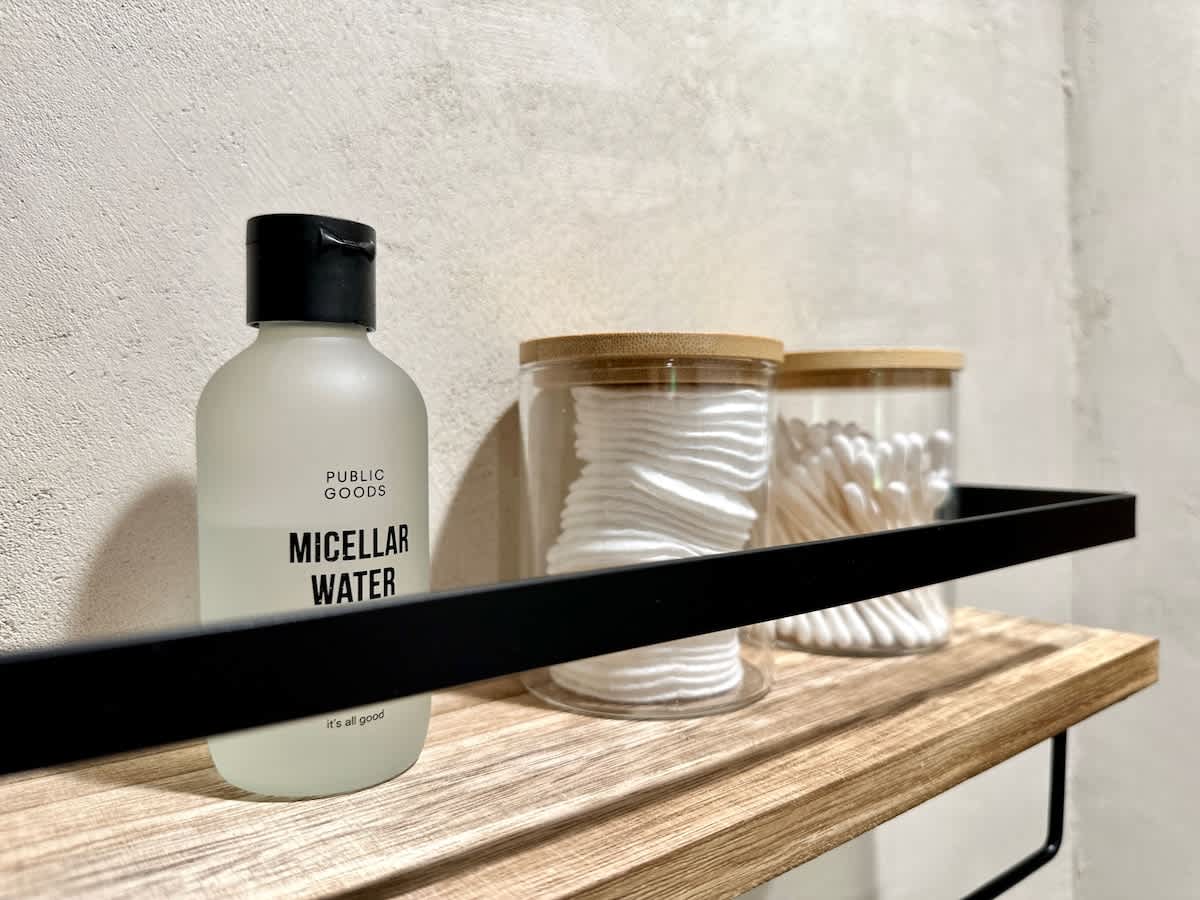 A close-up of a bathroom shelf featuring a bottle of micellar water and two glass jars with bamboo lids, one holding cotton pads and the other cotton swabs, set against a light textured wall.