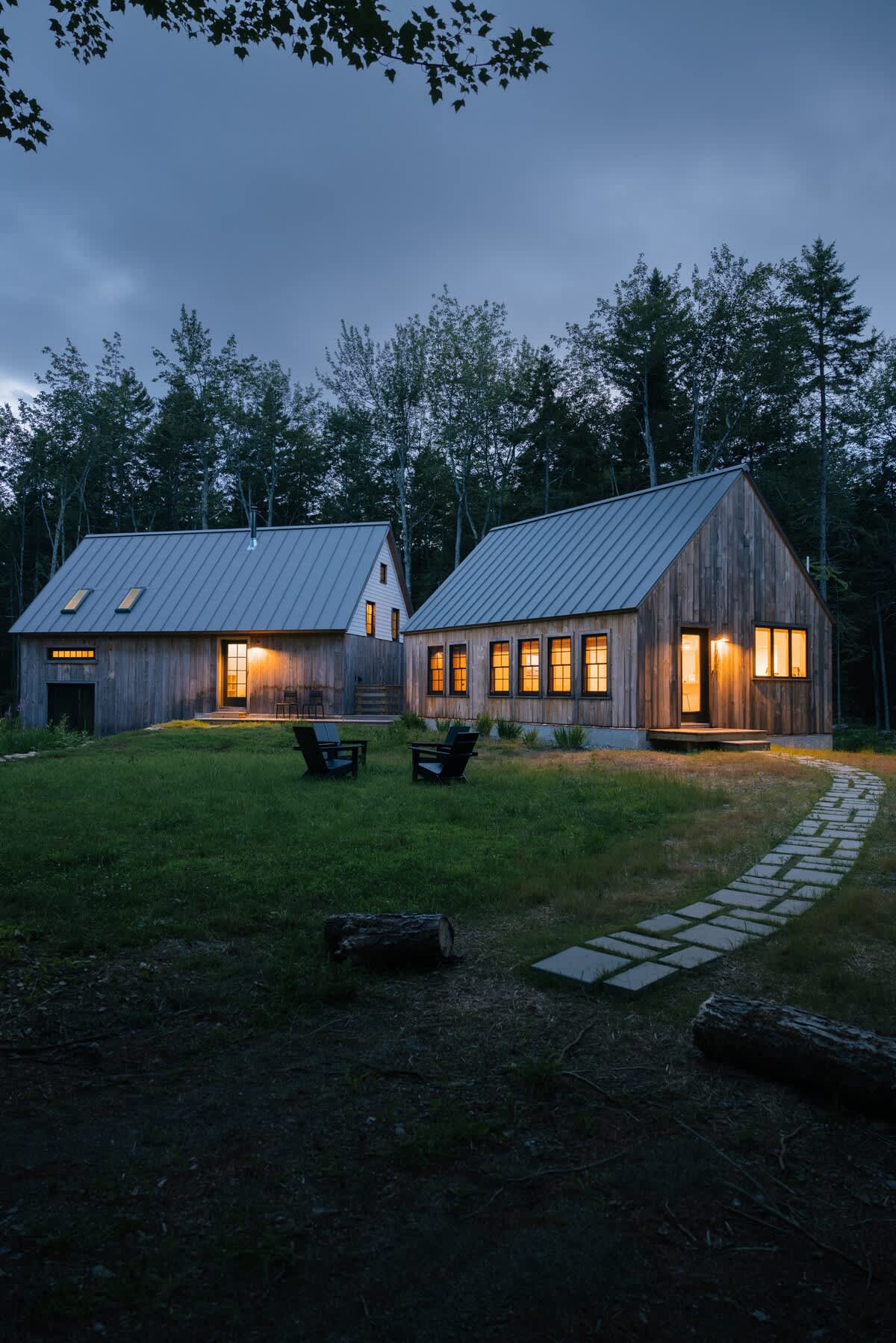 Two rustic wooden cabins with gray metal roofs and warm interior lights illuminate a grassy yard with a stone path at dusk. Adirondack chairs are arranged on the lawn.