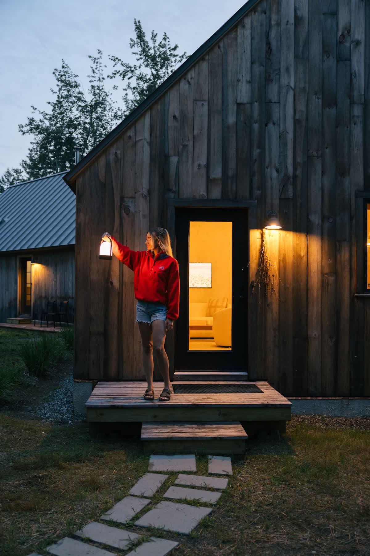 A woman in a red jacket stands on a wooden porch at dusk, holding a lit lantern, in front of a rustic wooden cabin with a dark door and warm interior light visible through the window.